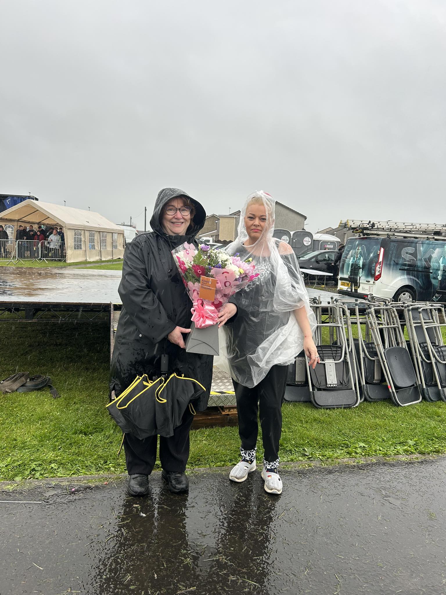 Clare Adamson MSP standing outdoors in the rain wearing a rain poncho, holding a large bouquet of flowers wrapped in pink paper. Another individual stands beside her, also in a rain poncho. They are on wet pavement with grass and a stage in the background.