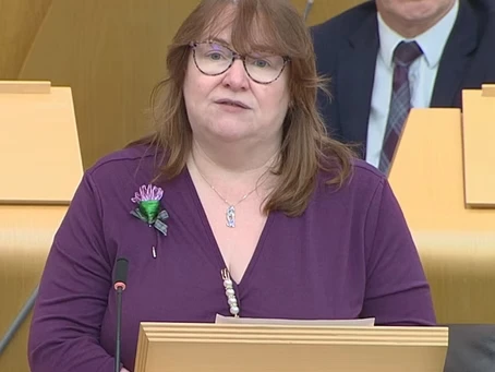 Clare Adamson MSP speaking at the podium in the Scottish Parliament chambers, wearing a purple top and flower brooch.