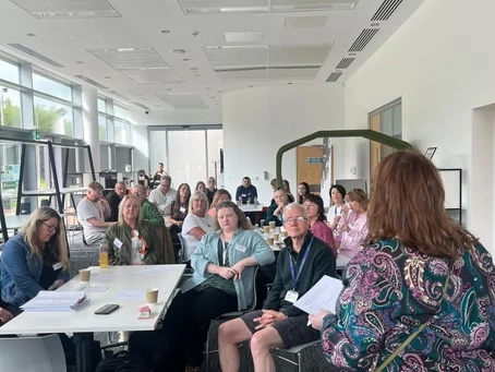 A group of people are seated in a bright, modern room with large windows. Clare Adamson MSP is standing at the front, addressing the attendees of the Community Action Network meeting in Motherwell and Wishaw. The attendees are listening attentively, with some taking notes.